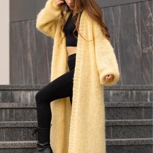 May include: A long, pale yellow open-front cardigan made of a fuzzy, textured knit material. The garment features long, loose sleeves with ribbed cuffs and a straight, floor-length hemline, worn over a black crop top and black leggings.