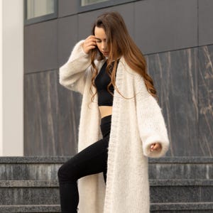 May include: A long, cream-colored, open-front knit cardigan with long sleeves and a textured, chunky yarn construction. The garment is worn over a black crop top and black leggings, featuring a ribbed trim along the front opening and cuffs.