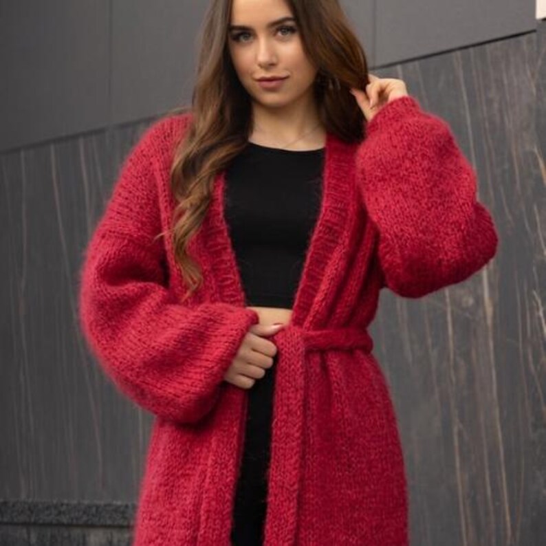May include: A vibrant red, chunky knit cardigan featuring long, voluminous sleeves and a matching tie belt at the waist. The garment is crafted from a thick, textured yarn with a fuzzy finish, designed with an open front and ribbed trim details.