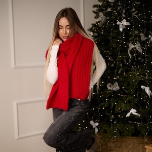 May include: A person wearing a bright red, chunky knit scarf. The scarf is draped over a white long-sleeve shirt and dark gray pants. A Christmas tree with white bows and lights is in the background.