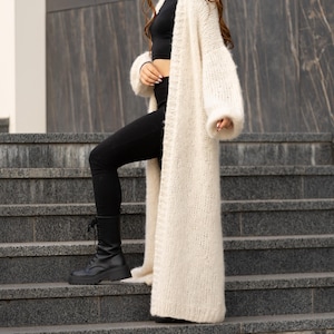 May include: A long, cream-colored, open-front knit cardigan with long sleeves, worn over a black crop top and black leggings, paired with black lace-up combat boots. The cardigan features a chunky, textured knit pattern and reaches mid-calf length.