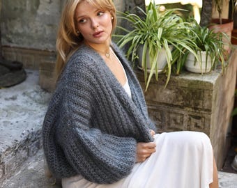 Peut inclure: Un gilet gris anthracite en tricot épais avec une texture côtelée, des manches longues et une ouverture sur le devant. Le vêtement est porté sur un haut blanc simple, présentant une texture douce et duveteuse ainsi qu'une coupe ample.
