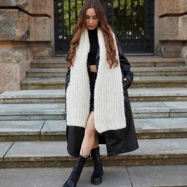 Puede incluir: Una bufanda larga de punto grueso en color crema con una textura suave y esponjosa. La bufanda se lleva sobre una parte superior negra de cuello alto, una gabardina negra de aspecto cuero y botas de combate negras con cordones.
