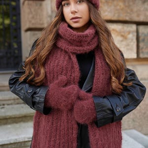 Puede incluir: Un conjunto de punto difuso en color granate oscuro que consta de un gorro, una bufanda larga y un par de manoplas. Los accesorios presentan una construcci&oacute;n de hilo grueso y texturizado, usados sobre un abrigo negro de manga larga.