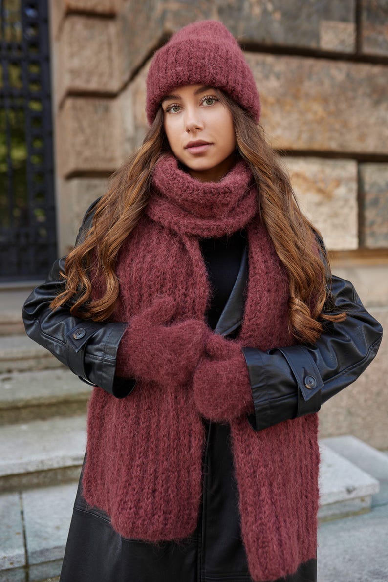 Puede incluir: Un conjunto de punto difuso en color granate oscuro que consta de un gorro, una bufanda larga y un par de manoplas. Los accesorios presentan una construcci&oacute;n de hilo grueso y texturizado, usados sobre un abrigo negro de manga larga.