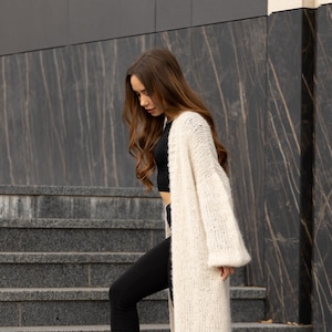 May include: A long, open-front cream-colored knit cardigan with a chunky texture and loose, wide sleeves. The garment is worn over a black cropped top and black leggings, featuring a soft, fuzzy yarn construction and a straight, relaxed fit.