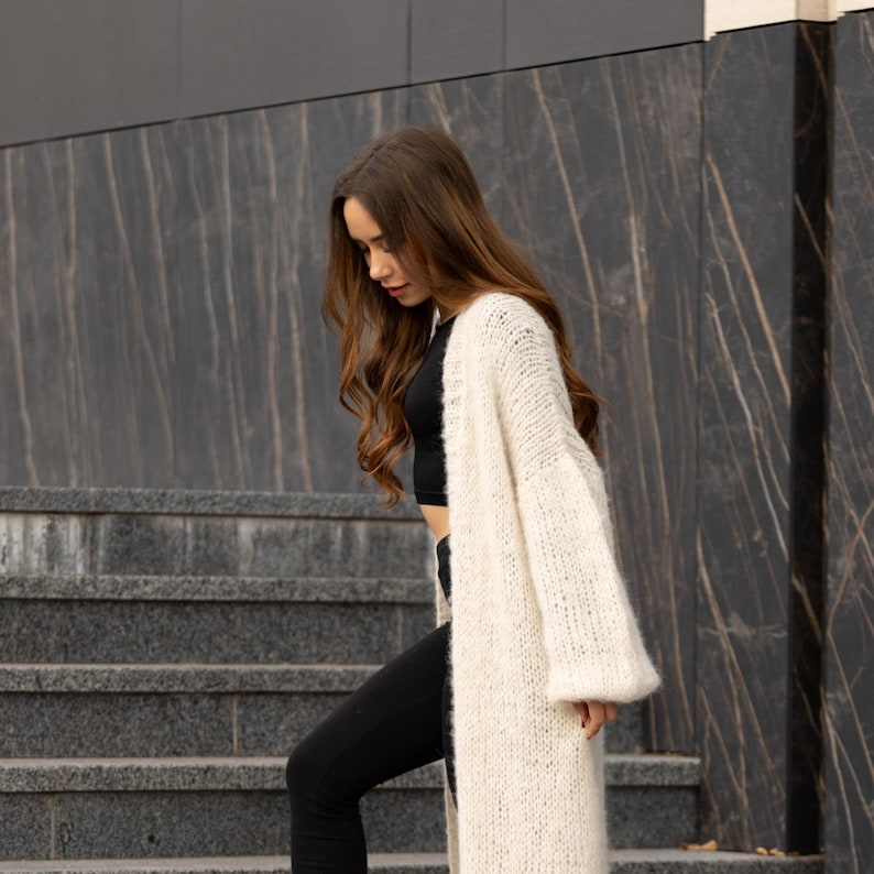 May include: A long, open-front cream-colored knit cardigan with a chunky texture and loose, wide sleeves. The garment is worn over a black cropped top and black leggings, featuring a soft, fuzzy yarn construction and a straight, relaxed fit.