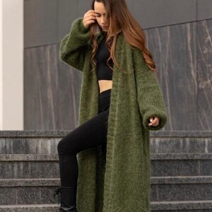 May include: A long, open-front olive green cardigan made of a chunky, fuzzy knit material. The garment features long, loose sleeves with ribbed cuffs and a straight, floor-length hemline, worn over a black crop top and black leggings.