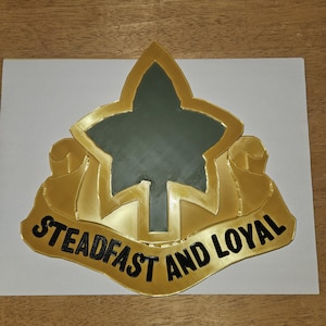 May include: A gold-colored emblem with a central green shape and the words "STEADFAST AND LOYAL" in black lettering. The emblem is set against a white background.