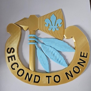 Pode incluir: Um emblema dourado com o texto "SECOND TO NONE" em preto. O emblema apresenta um tomahawk, uma flor-de-lis e três folhas azuis. O design está dentro de uma forma de ferradura.
