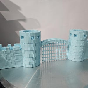May include: A light blue model castle with two cylindrical towers, a gate, and connecting walls. The castle is made of brick-like blocks and sits on a silver surface. The towers have square openings.