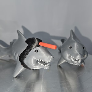 May include: Two gray 3D printed shark figurines. One shark has a black hat and an orange stick on top of its head. The other shark has a more traditional shark appearance.
