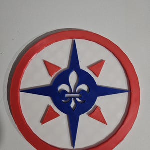 May include: A round, white emblem with a red border. A blue compass rose design is in the center, with a fleur-de-lis and red triangles. The emblem is likely a decorative item or a logo.