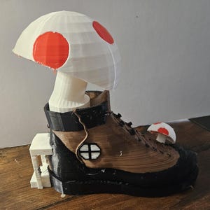 Puede incluir: Escultura impresa en 3D que presenta un hongo blanco con lunares rojos sobre una bota de senderismo marrón y negra. Un hongo más pequeño se encuentra en la bota. La escultura está sobre una base blanca.