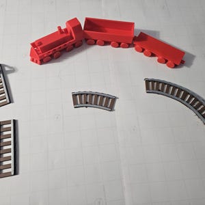 Puede incluir: Un tren de juguete rojo con tres vagones y tramos de vía. El tren es de plástico y está diseñado para niños. La vía incluye tramos rectos y curvos, y una escalera.
