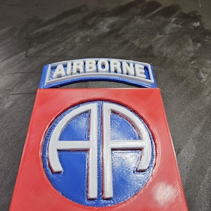 May include: A 3D-printed emblem with the word "AIRBORNE" in white on a blue background. Below, a red shield shape contains a blue circle with white "AA" letters. The emblem is likely a military or commemorative item.