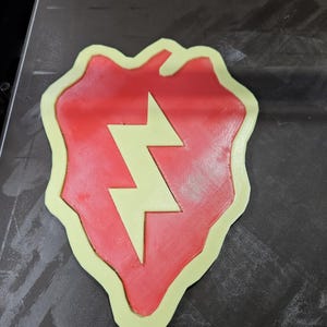May include: A 3D-printed emblem featuring a red shield shape with a yellow lightning bolt in the center. The emblem has a yellow border. The object is likely a military or organizational insignia.