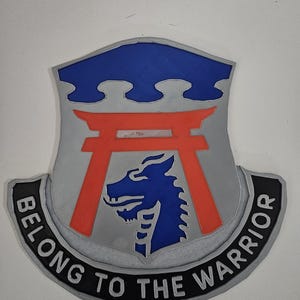 May include: A shield-shaped emblem with a blue top section and a red gate design. A blue dragon-like creature is centered, with the words "BELONG TO THE WARRIOR" in white on a black banner below. The emblem is gray and outlined in black.