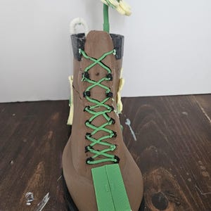 Puede incluir: Una bota marrón con cordones verdes y un rectángulo verde en la base de los cordones. Una flor amarilla y verde se coloca dentro de la bota. La bota está sobre una superficie marrón oscuro.