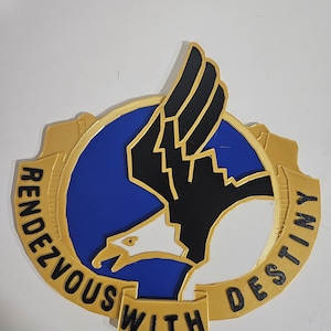 May include: A gold, blue, black, and white emblem with the words "Rendezvous With Destiny." The emblem features an eagle with outstretched wings, set against a blue background. The text is in a bold, sans-serif font.
