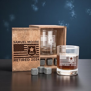 May include: A wooden gift box with two engraved whiskey glasses and whiskey stones. The glasses and box feature the text "SAMUEL MOORE", a police badge, an American flag design, and "RETIRED 2024". The glasses are clear and filled with a brown liquid.