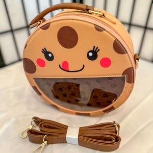Puede incluir: Un bolso redondo de color marrón y beige con forma de galleta con una ventana transparente de plástico que muestra un forro estampado en marrón y blanco. El bolso tiene una correa marrón y un asa de color dorado. La parte delantera del bolso tiene una cara de dibujos animados con ojos marrones, una boca roja y una lengua rosa.