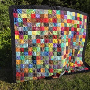 May include: A colourful patchwork quilt with a black border is displayed outdoors. The quilt features a vibrant array of squares in various colours, including red, yellow, blue, and green. The quilt is displayed on a grassy surface with trees in the background.