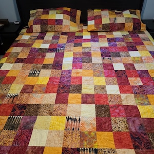 Puede incluir: Un edredón de retazos y dos fundas de almohada a juego en una variedad de colores cálidos, incluyendo rojo, naranja, amarillo y morado. El edredón presenta un patrón cuadrado con diferentes diseños de tela, creando un aspecto vibrante y texturizado. El marco de la cama es negro.