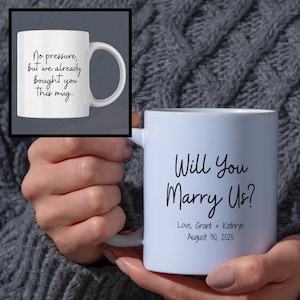 May include: White ceramic mug with the text "Will You Marry Us?" in black script. The mug also includes the names "Love, Grant + Kathryn" and the date "August 30, 2025". A smaller image shows a mug with the text "No pressure, but we already bought you this mug..."