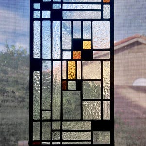 Op de afbeelding: Rechthoekig glas-in-loodpaneel met een geometrisch ontwerp. Het paneel heeft een zwart frame en diverse glasvormen in heldere, getextureerde en amberkleurige tinten. Het hangt aan een ketting, met uitzicht op de lucht en een huis.