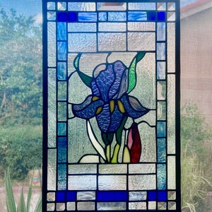 May include: Stained glass window panel featuring a detailed iris flower in shades of blue, green, and red. The flower is centered within a rectangular frame with a grid pattern of clear and blue glass. The panel is designed to hang.
