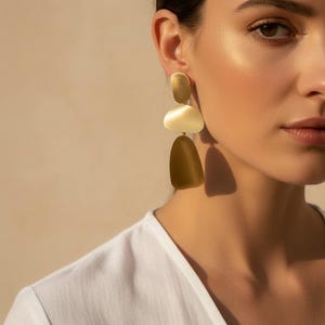 May include: A pair of gold-toned drop earrings featuring three stacked, smooth, organic-shaped metal plates. The earrings consist of a small oval top piece, a medium semi-circular middle piece, and a larger, elongated bottom piece in a matte finish.
