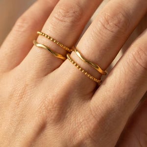 Op de afbeelding: Een set van vier goudkleurige ringen gedragen aan twee vingers. Twee ringen hebben een glad, golvend bandontwerp, terwijl de andere twee ringen bestaan uit een delicate, kralenachtige textuur. De ringen zijn per paar gestapeld op elke vinger.