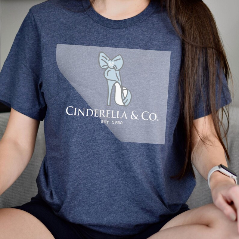 May include: Navy blue t-shirt featuring a light blue high-heeled shoe design with a bow and the text "Cinderella & Co. Est 1950." The design is on a light gray background. The shirt is made of a soft material.