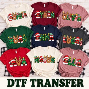 May include: A collection of nine short-sleeved t-shirts in various colors, each featuring a custom name printed in a festive Christmas-themed font decorated with reindeer antlers, Santa hats, elf hats, and colorful holiday lights.