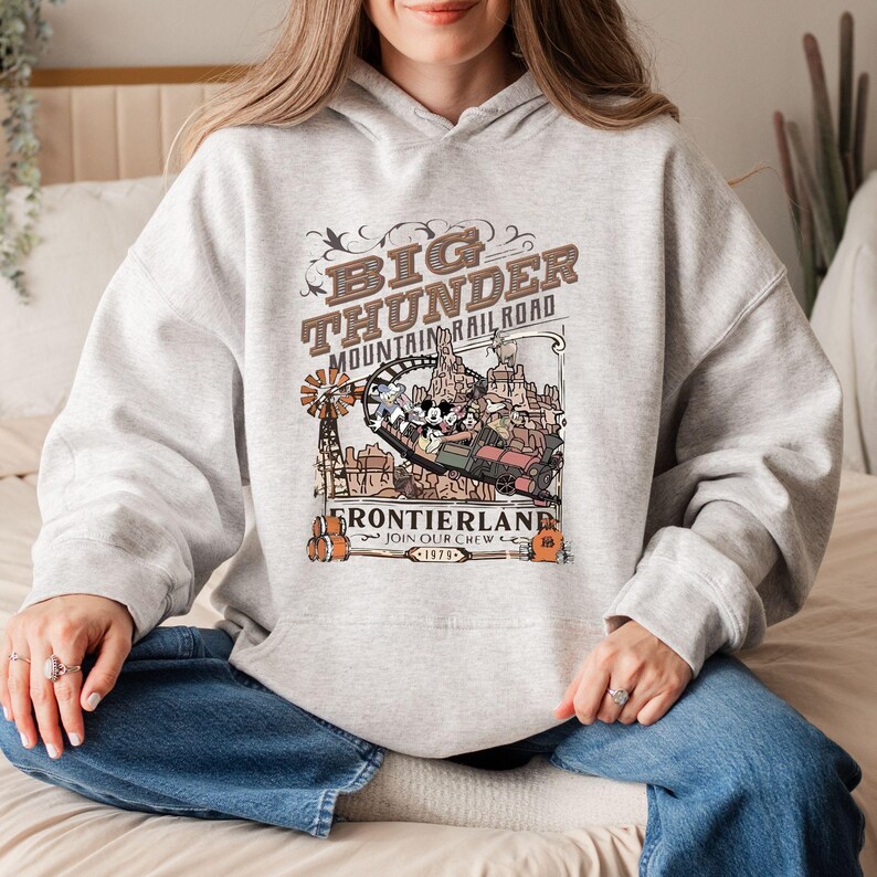 Peut inclure: Un sweat-shirt gris clair avec un motif du Big Thunder Mountain Railroad. Le design comprend le texte "Big Thunder Mountain Railroad" et "Frontierland" avec une illustration de style vintage du man&egrave;ge et des personnages.