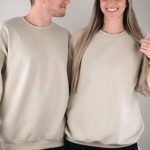 May include: Two people wearing matching beige crewneck sweatshirts. The sweatshirts have long sleeves and a relaxed fit. The fabric appears to be a soft, neutral color, suitable for casual wear. The image is well-lit, highlighting the texture of the material.