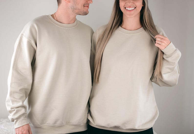 May include: Two people wearing matching beige crewneck sweatshirts. The sweatshirts have long sleeves and a relaxed fit. The fabric appears to be a soft, neutral color, suitable for casual wear. The image is well-lit, highlighting the texture of the material.