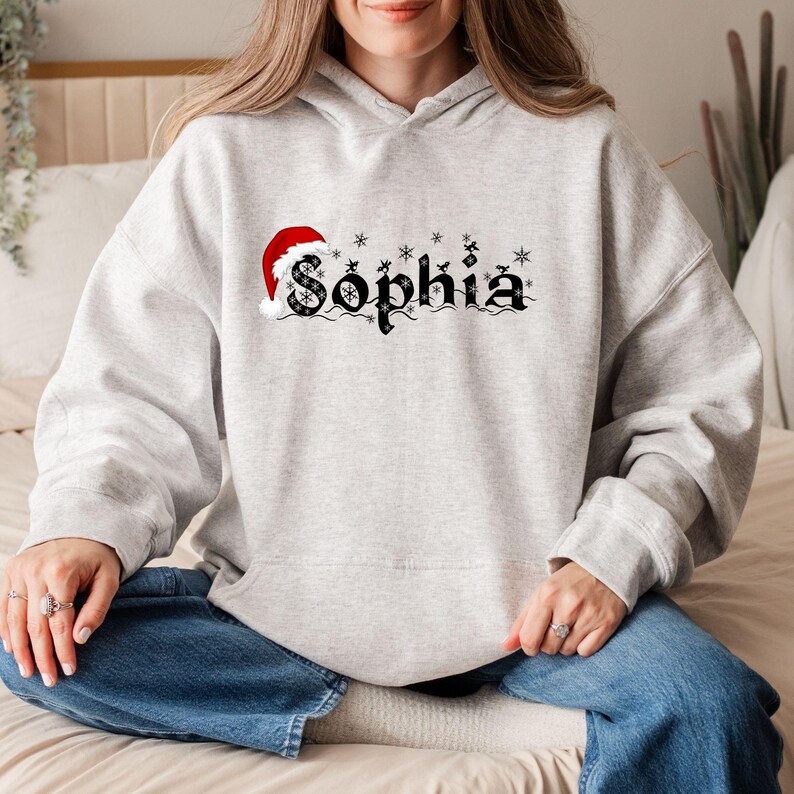 May include: A light gray hoodie with the name "Sophia" in black gothic lettering, adorned with snowflakes and a Santa hat. The hoodie is a comfortable, casual style, perfect for the holiday season.
