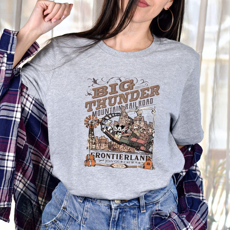 Peut inclure: T-shirt gris avec un motif du man&egrave;ge Big Thunder Mountain Railroad. Le motif comprend le texte "Big Thunder Mountain Railroad" et "Frontierland" avec une illustration de style vintage du man&egrave;ge et l'ann&eacute;e 1979.
