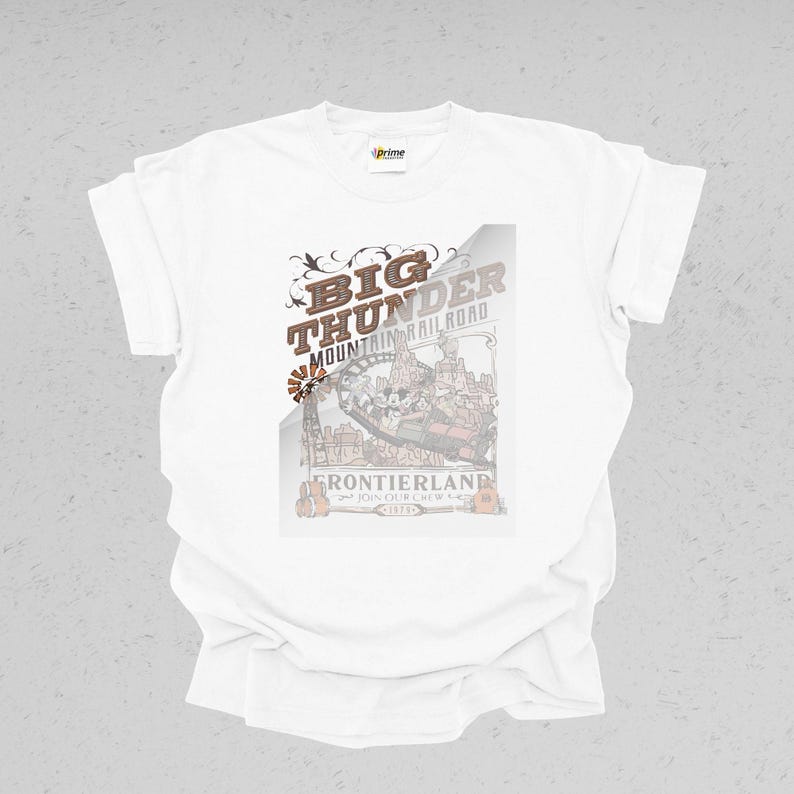 Peut inclure: T-shirt blanc avec un motif graphique pour Big Thunder Mountain Railroad. Le motif comprend le texte "Big Thunder Mountain Railroad" et "Frontierland". Le graphisme est dans des tons bruns et beiges.