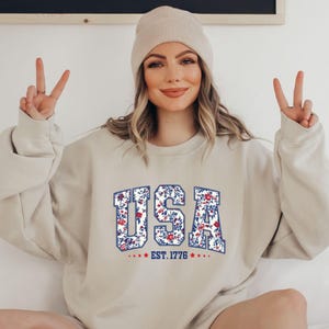 May include: Beige sweatshirt with the word "USA" in a floral pattern, outlined in blue. The text "EST. 1776" is below the word "USA". The sweatshirt is a neutral color and has a crew neck.