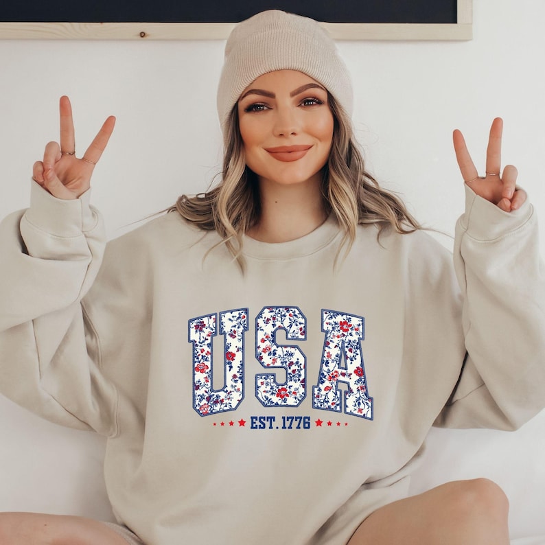 May include: Beige sweatshirt with the word "USA" in a floral pattern, outlined in blue. The text "EST. 1776" is below the word "USA". The sweatshirt is a neutral color and has a crew neck.