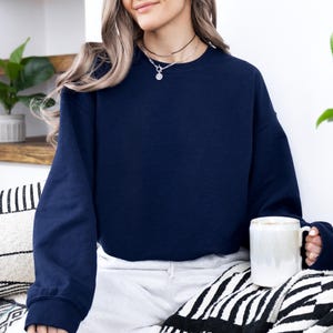 May include: A navy blue sweatshirt with long sleeves. The person is wearing a black necklace with a pendant. A white mug is held in the hand. The person is sitting on a black and white patterned blanket.