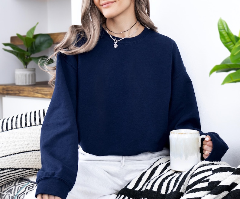 May include: A navy blue sweatshirt with long sleeves. The person is wearing a black necklace with a pendant. A white mug is held in the hand. The person is sitting on a black and white patterned blanket.
