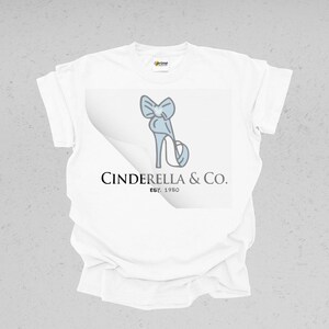May include: White t-shirt featuring a light blue high-heeled shoe with a bow design. The text "Cinderella & Co. EST. 1950" is printed below the shoe graphic. The shirt has a classic crew neck and short sleeves.