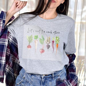 K&ouml;nnte beinhalten: Graues T-Shirt mit einer Aquarell-Illustration verschiedener Wurzelgem&uuml;se und dem Text "Let's root for each other".