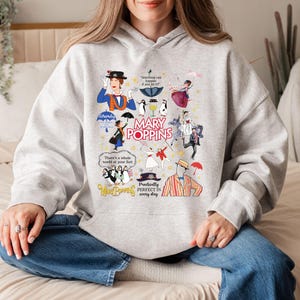 Puede incluir: Una sudadera con capucha gris claro con un dise&ntilde;o gr&aacute;fico colorido inspirado en la pel&iacute;cula "Mary Poppins". El dise&ntilde;o incluye ilustraciones de personajes, citas y el t&iacute;tulo de la pel&iacute;cula. La sudadera es una prenda c&oacute;moda e informal.