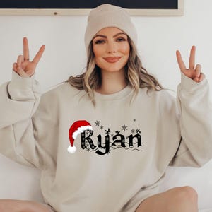 May include: Beige sweatshirt with the name "Ryan" in black gothic font, a red Santa hat, and snowflakes. The sweatshirt is a neutral color, suitable for the holiday season. The design is centered on the front.