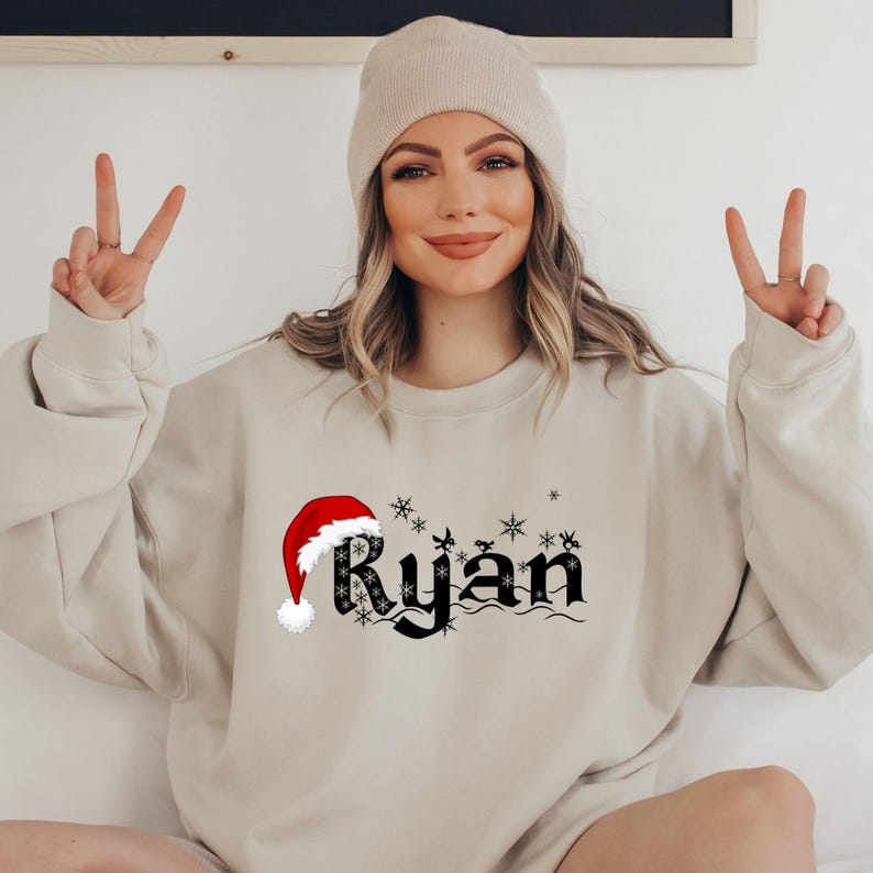 May include: Beige sweatshirt with the name "Ryan" in black gothic font, a red Santa hat, and snowflakes. The sweatshirt is a neutral color, suitable for the holiday season. The design is centered on the front.